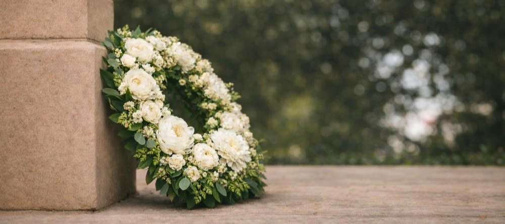 En vakker krans med ulike blomster og grønne blader, plassert ved en steinmur. De jordnære tonene og detaljene i blomstene gir et naturlig og verdig uttrykk, med en uskarp bakgrunn som skaper dybde i bildet. Foto.