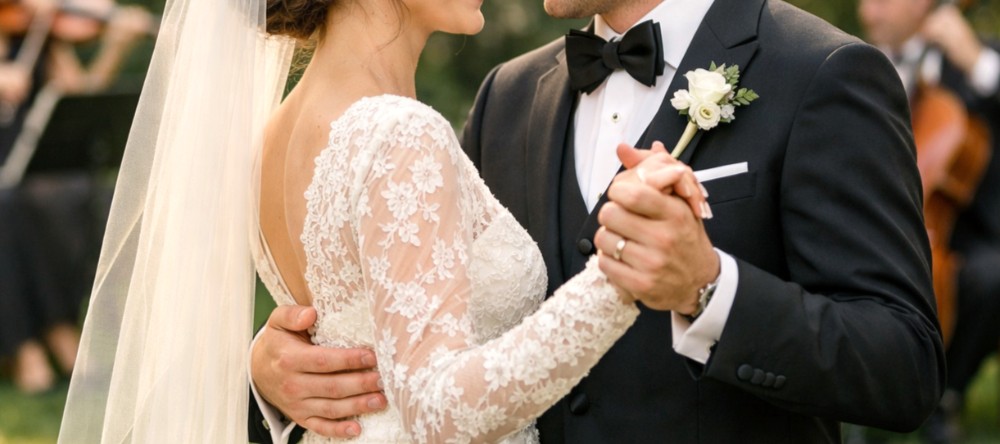 Et symbolsk bilde av et nygift par som holder hender, der begge bærer gifteringer. Bruden er kledd i en elegant, hvit kjole, mens brudgommen har på seg en klassisk, mørk dress. Bildet fokuserer på hendene og formidler en følelse av kjærlighet og samhold. Foto.